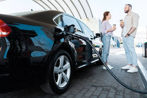 Selective focus of black car refueling with benzine near man and woman — Stock Photo