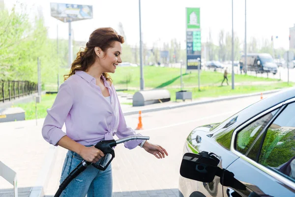 Mujer alegre sosteniendo la bomba de combustible cerca de coche negro en la gasolinera - foto de stock