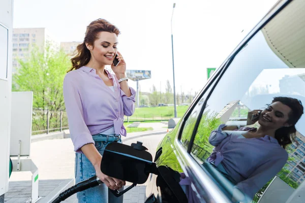 Femme heureuse tenant pompe à carburant tout en ravitaillant la voiture avec de la benzine et parler sur smartphone — Photo de stock