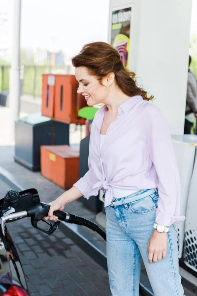 Mujer feliz sosteniendo la bomba de combustible mientras repostan automóvil con bencina en la gasolinera - foto de stock