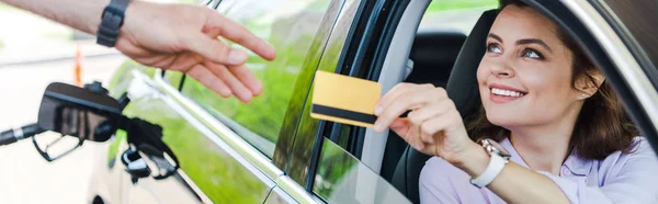 Panoramic shot of cheerful woman sitting in car and giving credit card to worker at gas station — Stock Photo