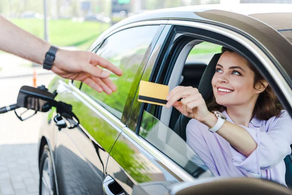 Foco seletivo da mulher feliz sentada no carro e dando cartão de crédito ao trabalhador no posto de gasolina — Fotografia de Stock