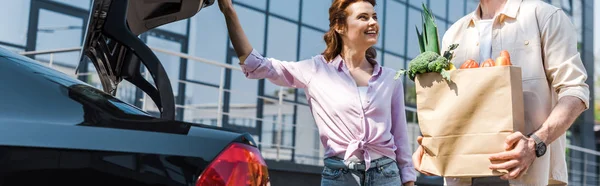 Panoramic shot of happy woman looking at man holding paper bag near car — Stock Photo