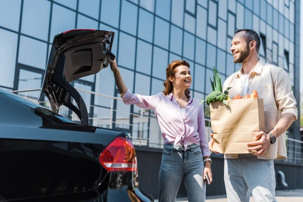 Vista basso angolo di donna felice guardando l'uomo con sacchetto di carta in piedi vicino auto — Foto stock