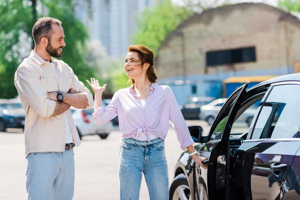 Happy woman gesturing while holding handle and opening car near handsome man — Stock Photo