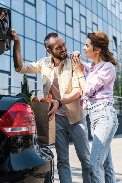 Happy bearded man putting paper bag in car trunk near attractive woman — Stock Photo