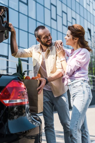 Hombre barbudo feliz poniendo bolsa de papel en el maletero del coche cerca de mujer atractiva - foto de stock