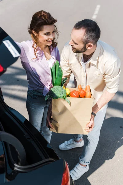 Overhead view of happy bearded man putting paper bag in car trunk near attractive woman — Stock Photo