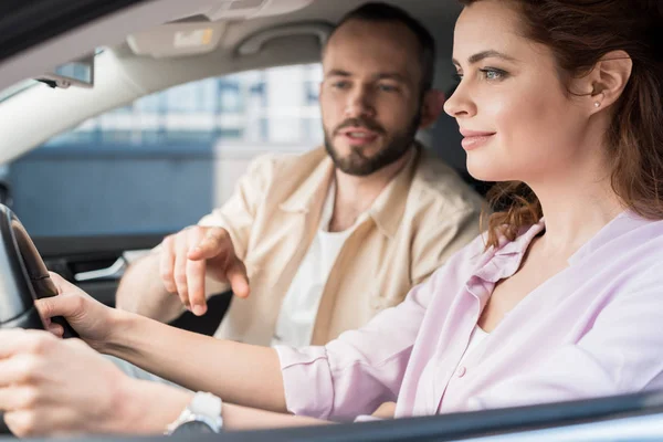 Selektiver Fokus eines gutaussehenden Mannes, der mit dem Finger auf das Lenkrad neben der glücklichen Frau im Auto zeigt — Stockfoto