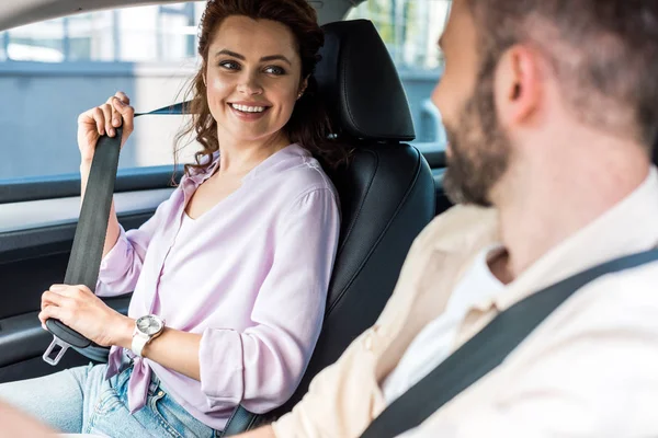 Вибірковий фокус привабливої і веселої жінки, що тримає ремінь безпеки і дивиться на чоловіка в машині — стокове фото