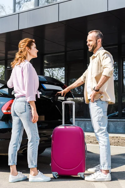 Handsome and cheerful man looking at happy woman while standing near car and pink luggage — Stock Photo