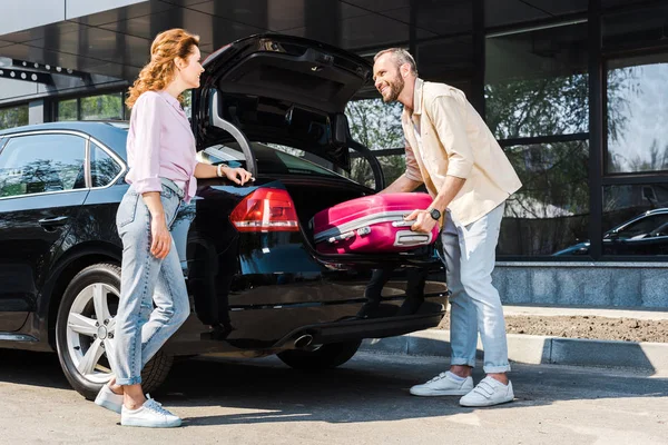Низкий угол зрения счастливого человека положить розовый багаж в багажник автомобиля рядом с женщиной — стоковое фото