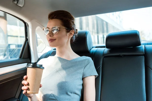 Cheerful businesswoman in glasses sitting in car and holding paper cup — Stock Photo