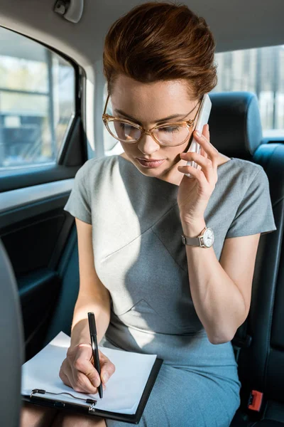 Attractive businesswoman in glasses writing while talking on smartphone in car — Stock Photo