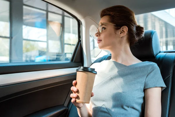Vista basso angolo di donna in bicchieri che tengono tazza di carta mentre seduto sulla macchina — Foto stock