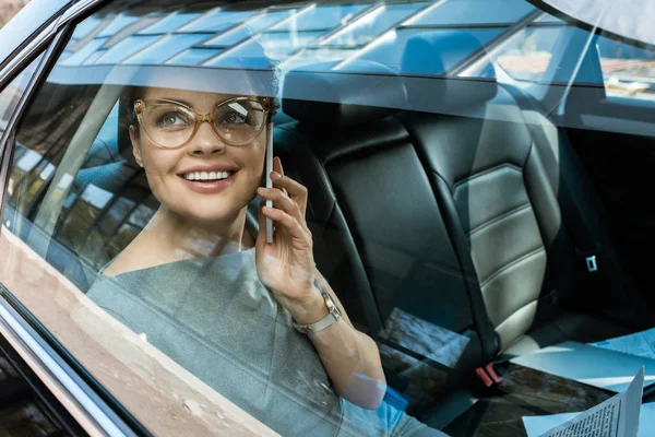 Happy businesswoman in glasses looking at window while talking on smartphone — Stock Photo