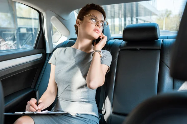 Selektive Fokussierung unzufriedener Geschäftsfrau in Brille, die im Auto mit Smartphone spricht — Stockfoto