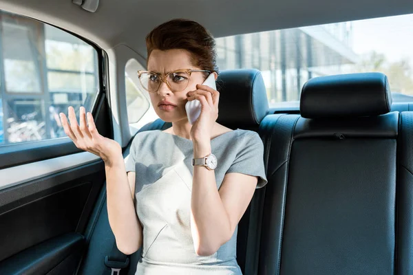 Displeased businesswoman in glasses talking on smartphone and gesturing in car — Stock Photo