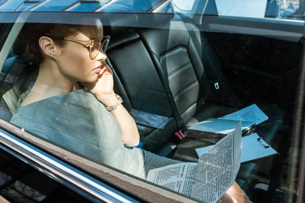 Attractive businesswoman in glasses reading newspaper while talking on smartphone in car — Stock Photo