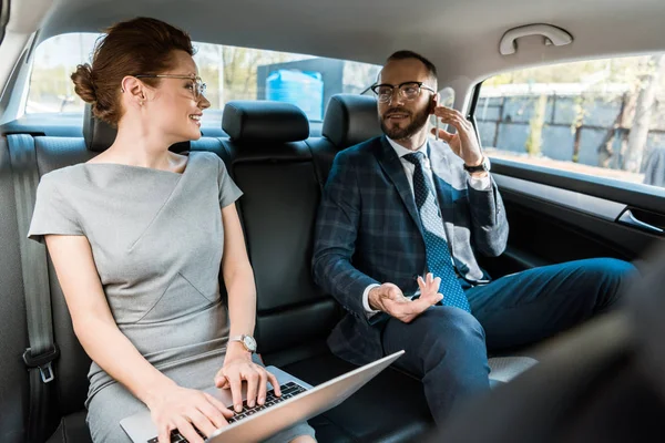 Selective focus of businessman in glasses talking on smartphone and looking at businesswoman typing on laptop in car — Stock Photo