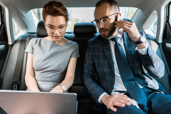 Hombre de negocios guapo mirando a la computadora portátil mientras habla en el teléfono inteligente cerca de la mujer de negocios - foto de stock