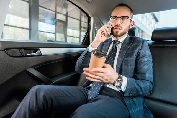 Low angle view of handsome businessman talking on smartphone and holding paper cup in car — Stock Photo