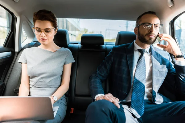 Привлекательная деловая женщина с ноутбуком рядом с бизнесвумен разговаривает на смартфоне в машине — стоковое фото
