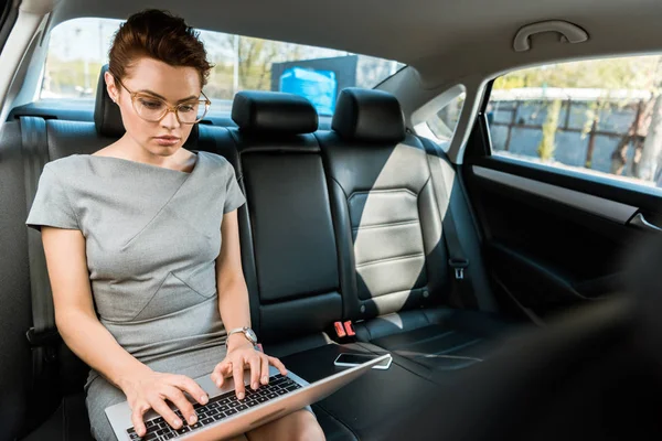 Foco seletivo de mulher de negócios bonita em óculos digitando no laptop no carro — Fotografia de Stock