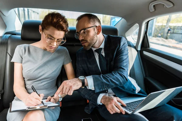 Schöner Geschäftsmann zeigt mit dem Finger auf Klemmbrett, während er mit Laptop neben Frau im Auto sitzt — Stockfoto