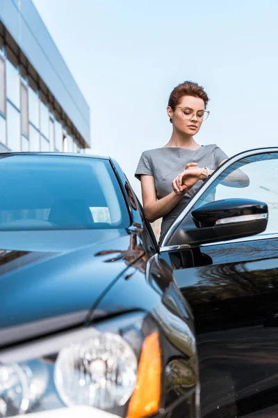 Selektiver Fokus einer Frau mit Brille, die auf die Uhr schaut, während sie in der Nähe eines schwarzen Autos steht — Stockfoto