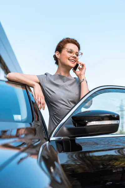 Vue à angle bas de femme heureuse parlant sur smartphone près de voiture noire — Photo de stock