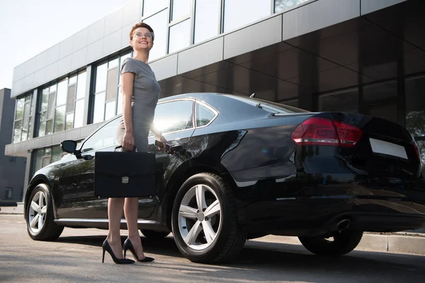 Visão de baixo ângulo de empresária feliz em pé perto de carro preto com pasta — Fotografia de Stock
