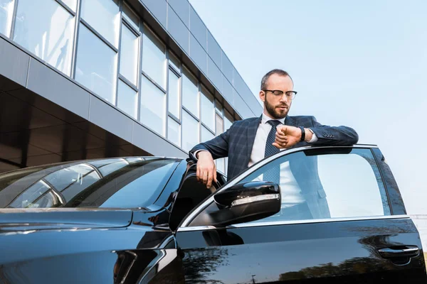 Vista de ángulo bajo de hombre de negocios en gafas mirando el reloj cerca de coche negro - foto de stock