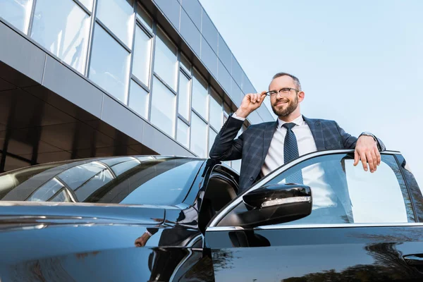 Low angle view of businessman in suit touching glasses while standing near black car — Stock Photo