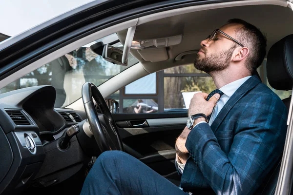 Low angle view of bearded businessman in glasses touching tie while sitting in car — Stock Photo