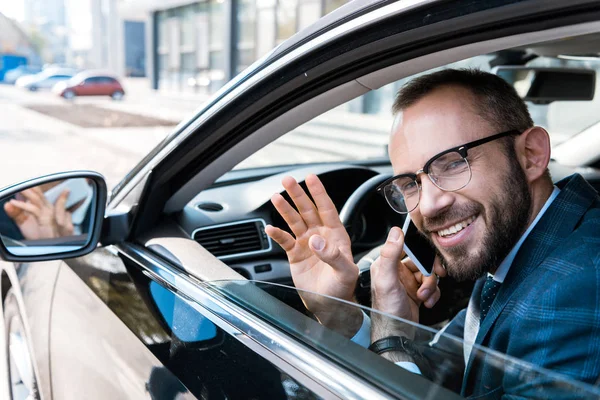 Selective focus of happy bearded businessman in glasses talking on smartphone while waving hand in car — Stock Photo