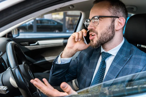 Foco seletivo de homem de negócios barbudo irritado falando no smartphone e gesticulando no carro — Fotografia de Stock