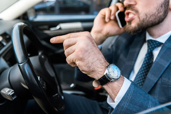 Enfoque selectivo de hombre de negocios barbudo enojado hablando en el teléfono inteligente y señalando con el dedo en el coche - foto de stock