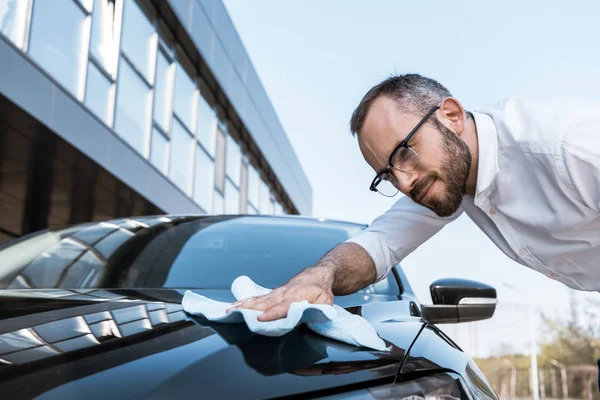 Vista de ángulo bajo de hombre de negocios feliz en gafas de limpieza coche negro con tela blanca cerca del edificio - foto de stock