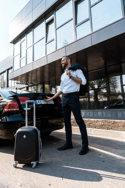 Vista de ángulo bajo de hombre de negocios feliz de pie cerca de coche negro con equipaje - foto de stock