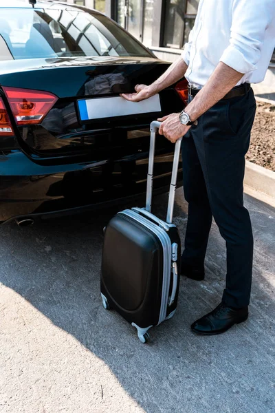 Обрезанный вид бизнесмена, открывающего багажник автомобиля, стоя с багажом — стоковое фото