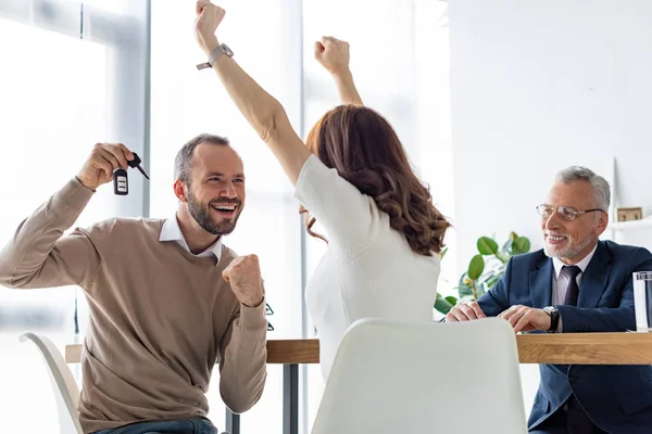 Selective focus of cheerful man holding car keys and gesturing near woman and car dealer — Stock Photo