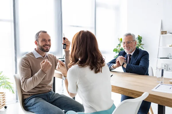 Enfoque selectivo de hombre feliz sosteniendo las llaves del coche y el gesto cerca de la mujer y concesionario de coches - foto de stock