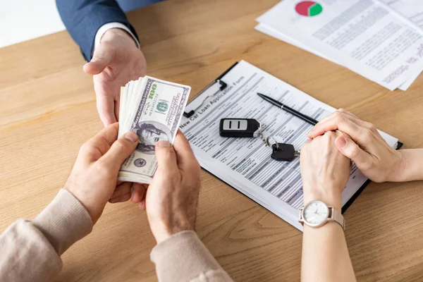 Vista ritagliata di uomo dando banconote dollaro al rivenditore di auto vicino alla donna con le mani serrate — Foto stock