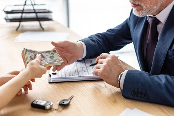 Vista recortada de la mujer que da billetes de dólar al concesionario de automóviles barbudos en la oficina - foto de stock