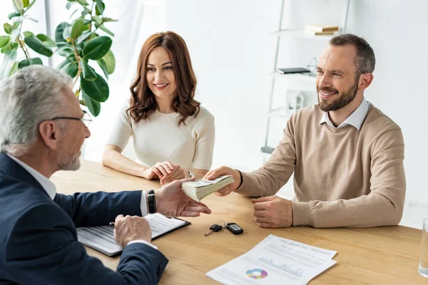 Cheerful man giving dollar banknotes to car dealer near attractive woman — Stock Photo