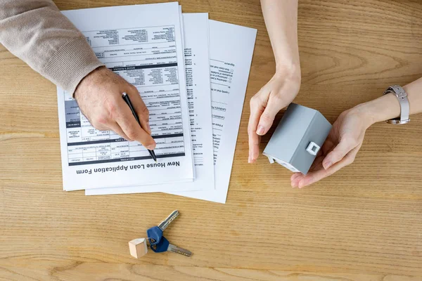 Top view of man holding pen near new house loan application form and woman with house model in hands — Stock Photo