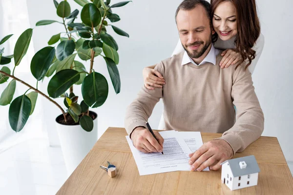 Schöner Mann unterschreibt Vertrag nahe glücklicher Frau, Schlüssel und Hausmodell — Stockfoto
