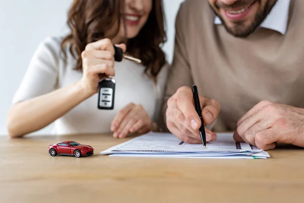 Enfoque selectivo del coche de juguete rojo cerca del hombre barbudo que firma el contrato y la mujer que sostiene la llave del coche - foto de stock