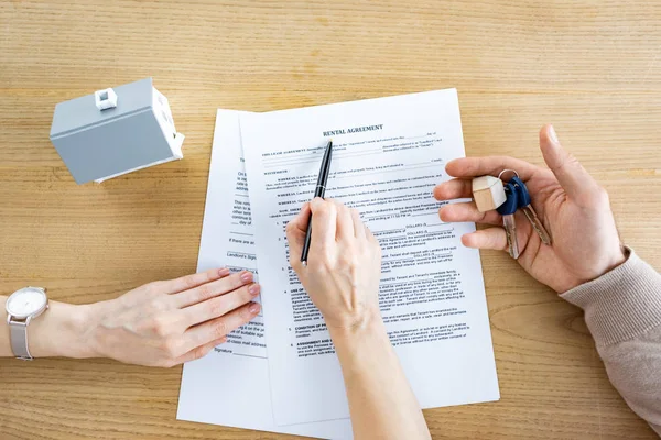 Vista superior da mulher segurando caneta perto do documento com lettering contrato de aluguel e homem com as chaves na mão — Fotografia de Stock